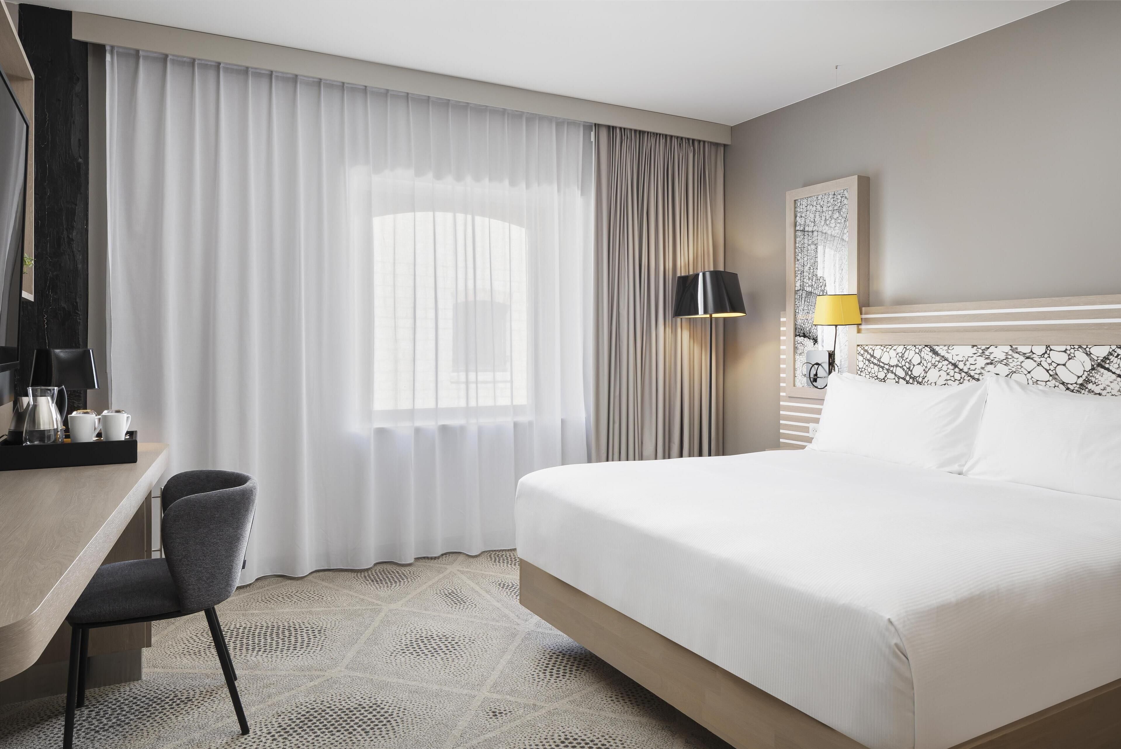 Una habitación de hotel con cama, escritorio, silla, lámpara y un amplio ventanal con cortinas.