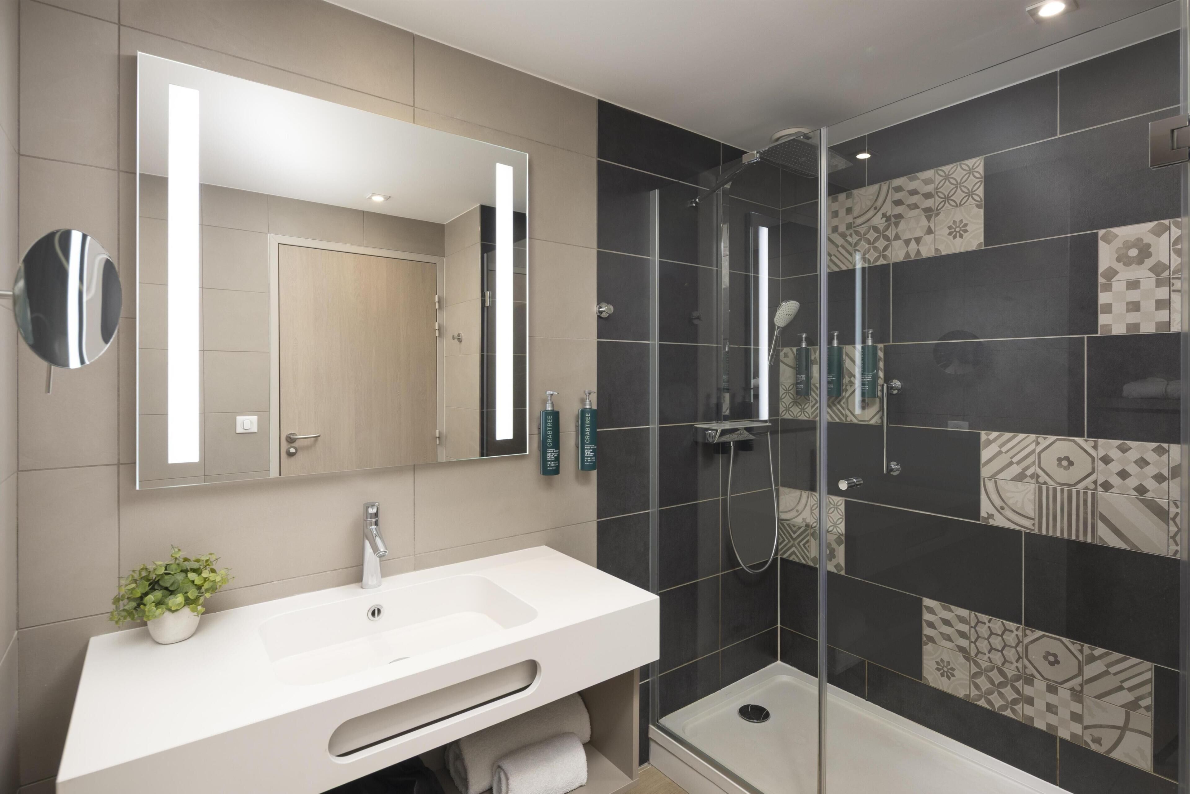 A modern bathroom with a large mirror, a white sink, a shower enclosure, and patterned tiles.