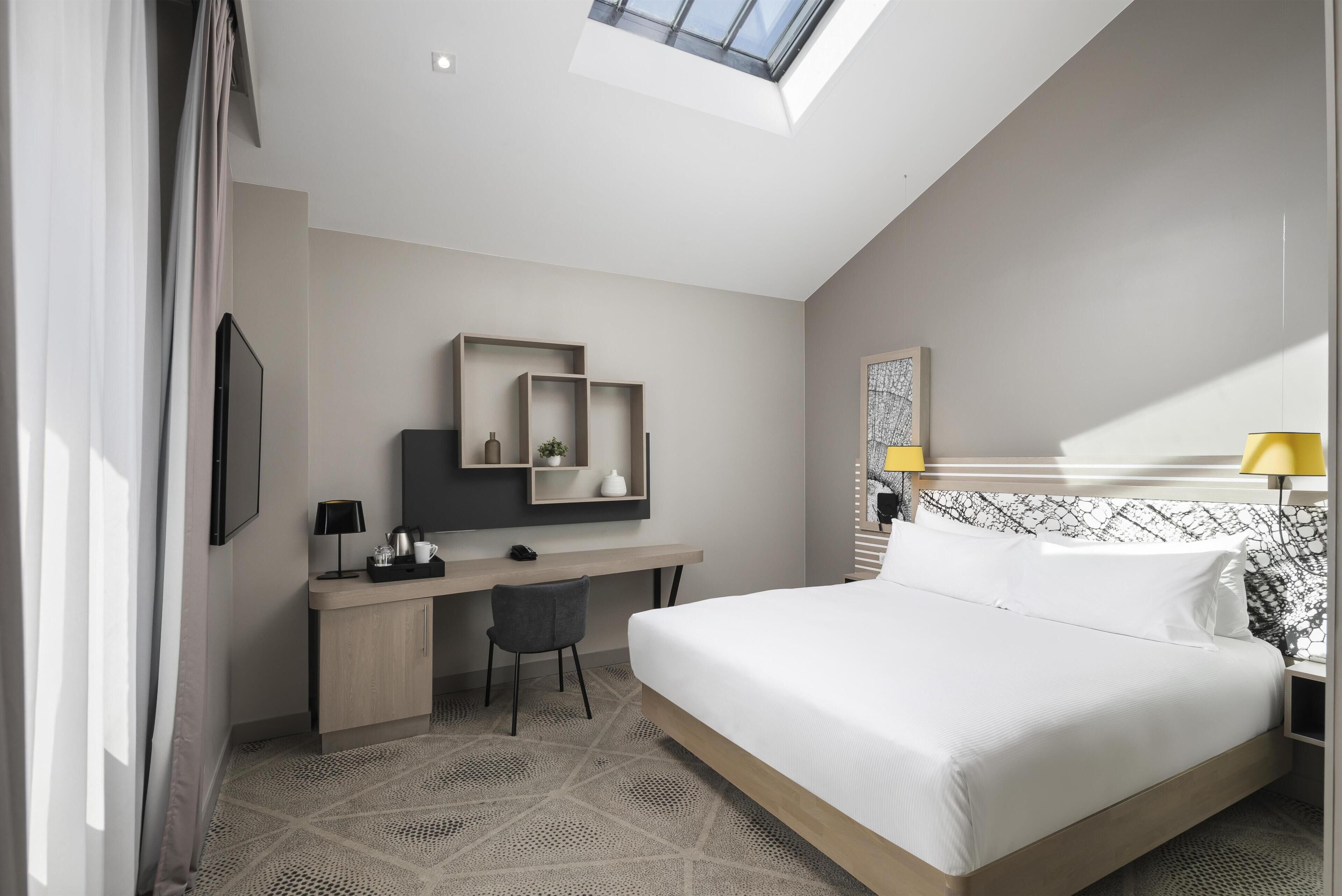 Una habitación de hotel moderna con tragaluz, una cama, un escritorio con silla y una mesita de noche con lámpara.
