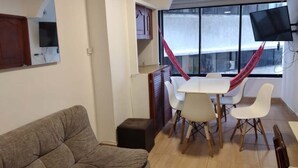 Een woonkamer met een bank, een eettafel met stoelen, een aan de muur bevestigde televisie en een raam met gordijnen.