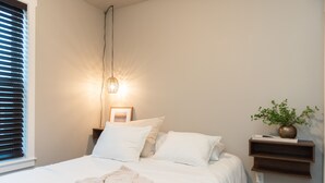 Een net opgemaakt bed met witte lakens en kussens, een bedlampje, een klein nachtkastje met een plantje, en een wandplank.