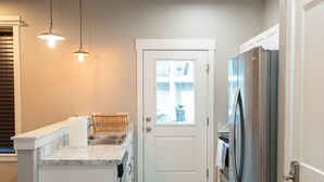 Een moderne keuken met een wit eiland, roestvrijstalen apparatuur, hanglampen en uitzicht door de deur.