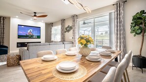 Un comedor moderno con una mesa de madera dispuesta para seis comensales, un televisor de pantalla plana y un ventanal con cortinas.