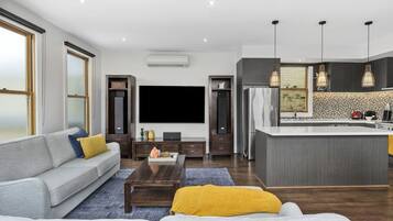 A modern living room with a sofa, coffee table, and TV. There's a kitchen area with a large island and pendant lights.