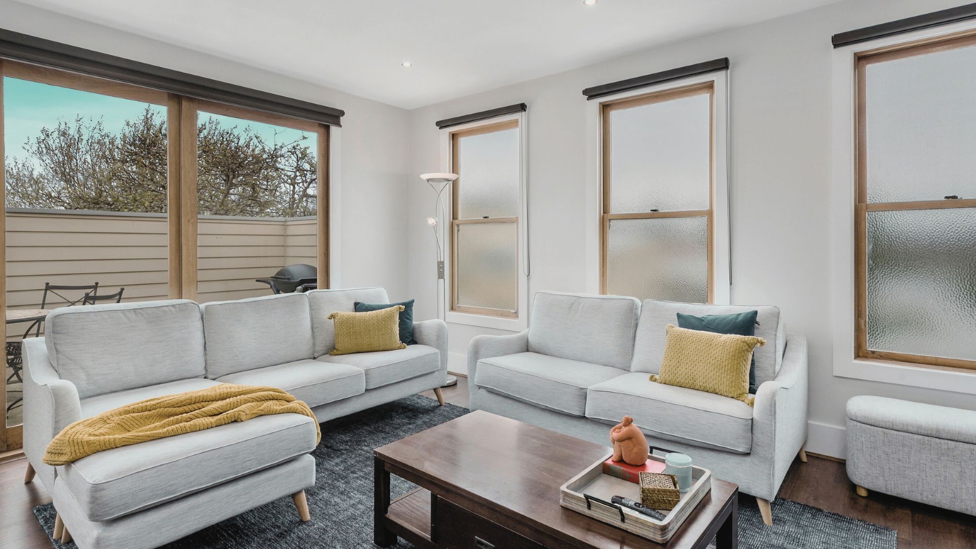 A modern living room with two grey sofas, a wooden coffee table, and large windows with blinds.