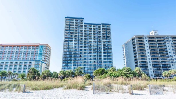 A beachfront hotel with multiple high-rise buildings, featuring balconies and a sandy beach in the foreground.