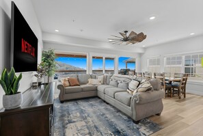 A modern living room with a sectional sofa, a flat-screen TV, a ceiling fan, and a view of the outdoors.