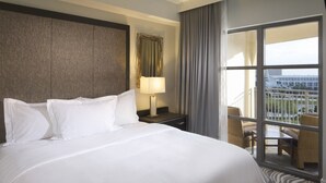 A hotel room with a large bed, a nightstand with a lamp, a view of the city through a balcony with wicker furniture.