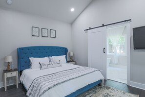 A bedroom with a blue headboard, white bedding, and a patterned throw. There are two bedside tables with lamps, a large window with a view, and a sliding door to a balcony.