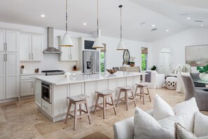 A modern kitchen with white cabinetry, a central island with stools, pendant lighting, and a large window.