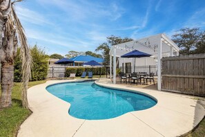 A swimming pool with a curved edge, surrounded by a wooden fence, and shaded by umbrellas. - Pelican Retreat by Avantstay 5 Mins TO Beach Pet Friendly w/ Pool & Bbq! (Destin)