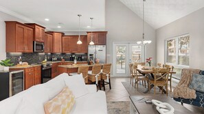 A modern kitchen with wooden cabinets, a dining area with a table and chairs, a sofa, and a large window with blinds.