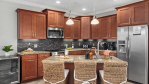 A modern kitchen with wooden cabinets, stainless steel appliances, and a central island with wicker chairs. - Pelican Retreat by Avantstay 5 Mins TO Beach Pet Friendly w/ Pool & Bbq! (Destin)