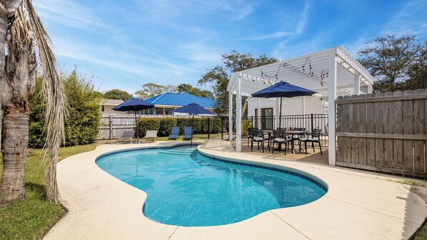 A swimming pool with a curved edge, surrounded by a patio area with outdoor furniture and umbrellas. - Pelican Retreat by Avantstay 5 Mins TO Beach Pet Friendly w/ Pool & Bbq! (Destin)