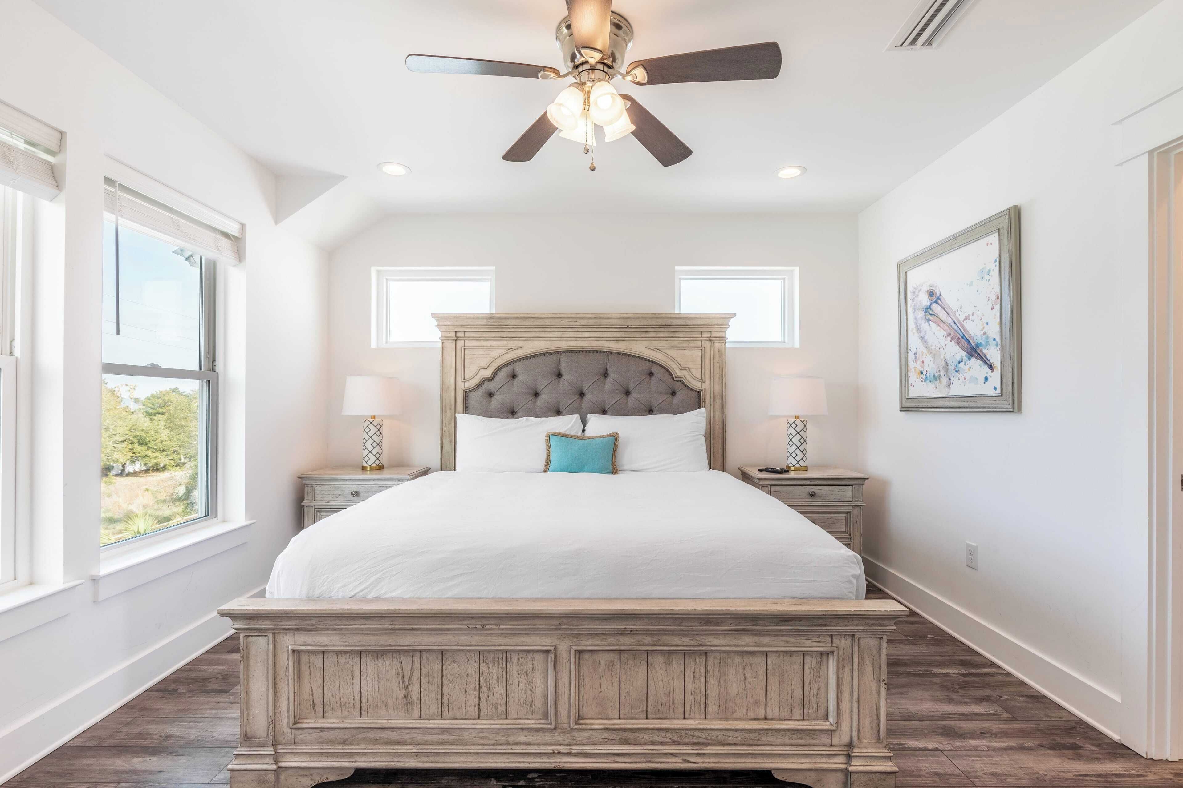 A bedroom with a large bed, a ceiling fan, two bedside tables with lamps, and a window providing a view of the outdoors.