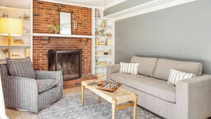 Una sala de estar con chimenea de ladrillo, un sofá gris, un sillón estampado y una mesa de centro tejida.