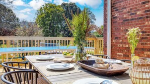 Un área de comedor al aire libre con una mesa de madera preparada para dos personas, rodeada de sillas, una mesa de ping pong y un muro de ladrillo.