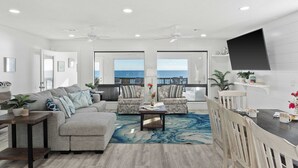 A modern living room with a large sofa, two armchairs, a coffee table, and a dining table with chairs. There is a large window with a view of the ocean. - Destiny on the Sand by Avantstay Direct Beach Access! Bbq on Large Ocean Facing Patio (Destin)