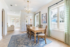 A dining area with a large wooden table, six chairs, a blue patterned rug, and a chandelier.