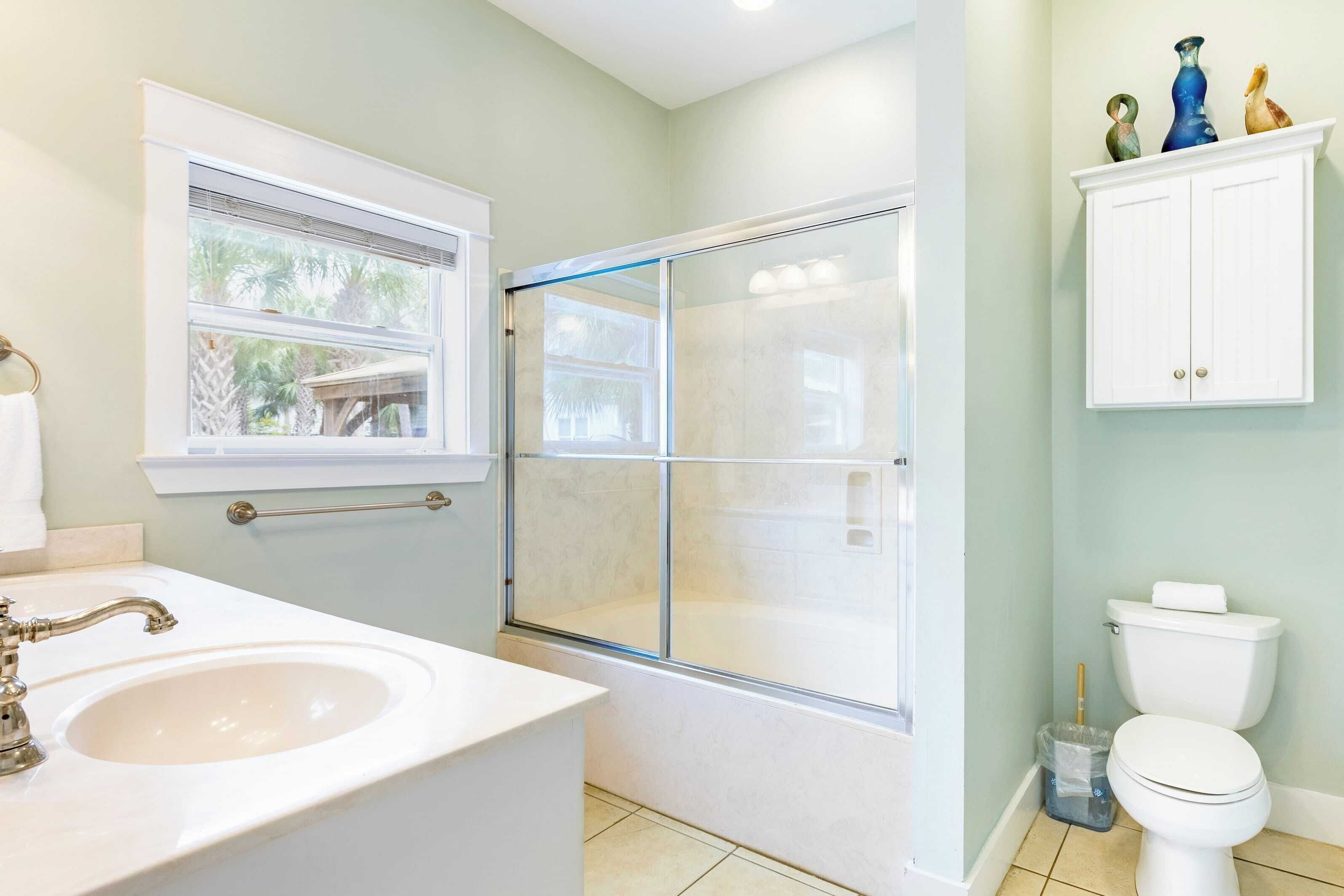 A bathroom with a glass-enclosed shower, a sink, a toilet, and a window with a view of palm trees.