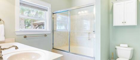 A bathroom with a glass-enclosed shower, a sink, a toilet, and a window with a view of palm trees.
