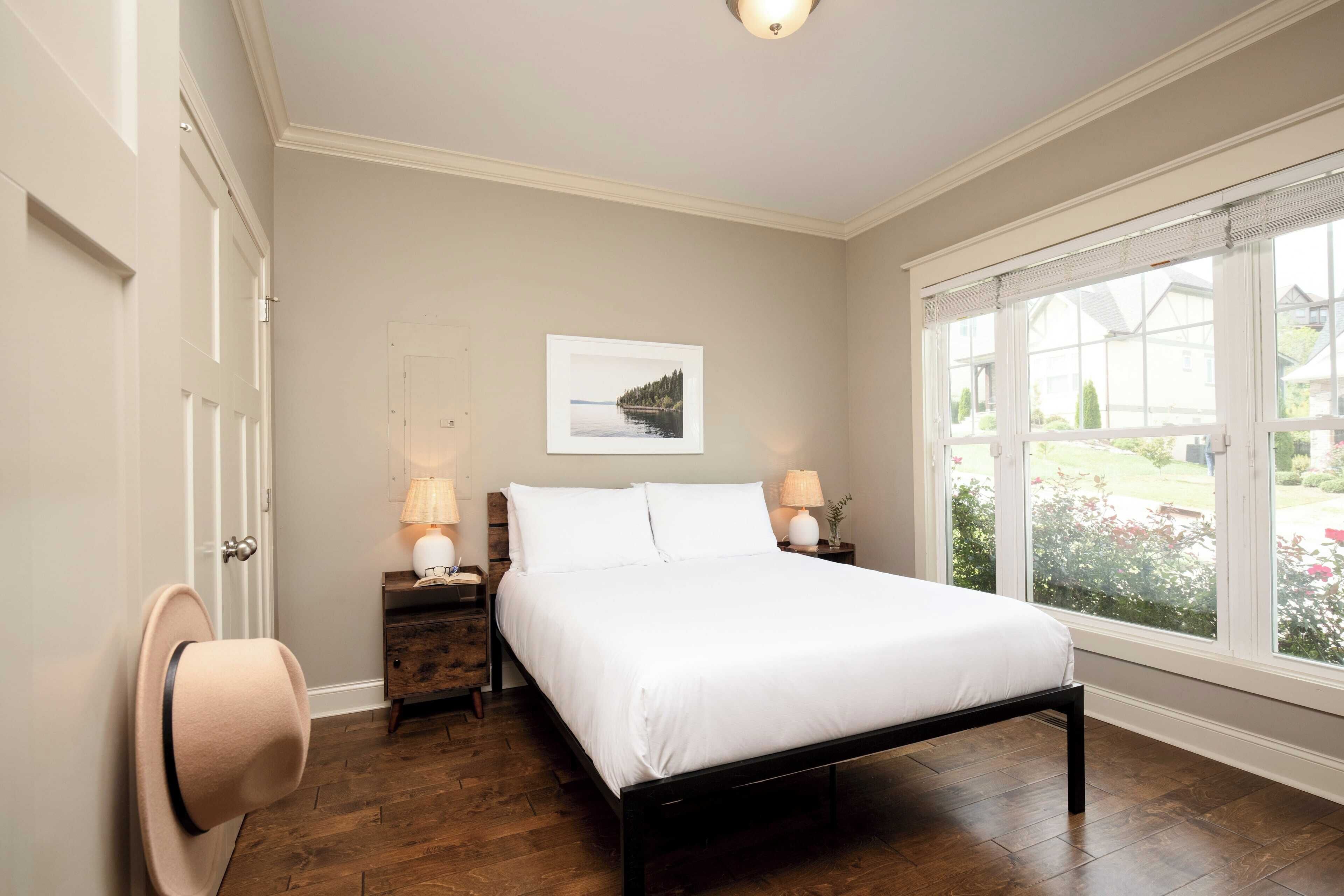 A bedroom with a bed, a nightstand, a lamp, a window with a view of greenery, and a door.