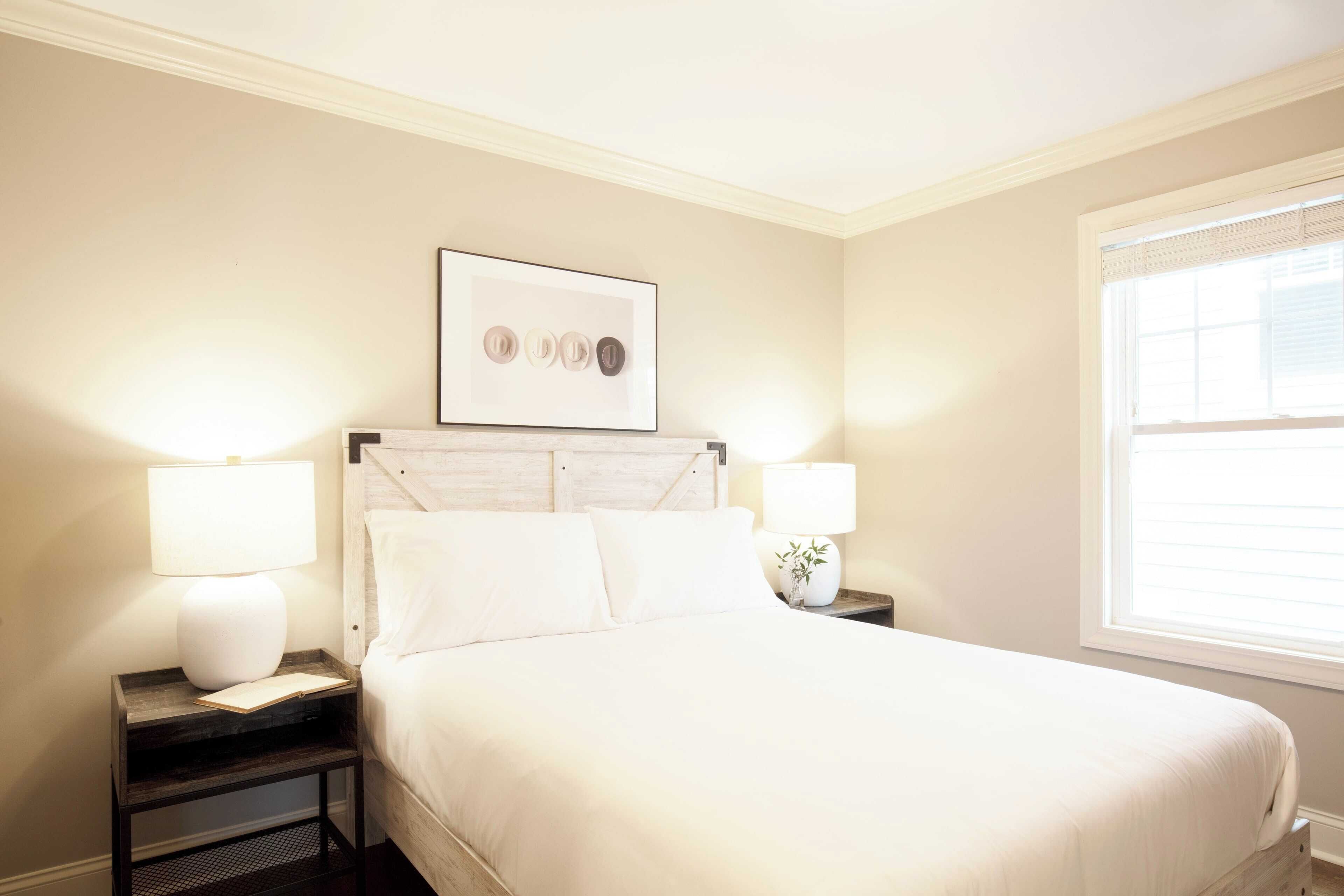 A neatly made bed with white linens, two bedside lamps, a framed picture above the bed, and a window with blinds to the right.