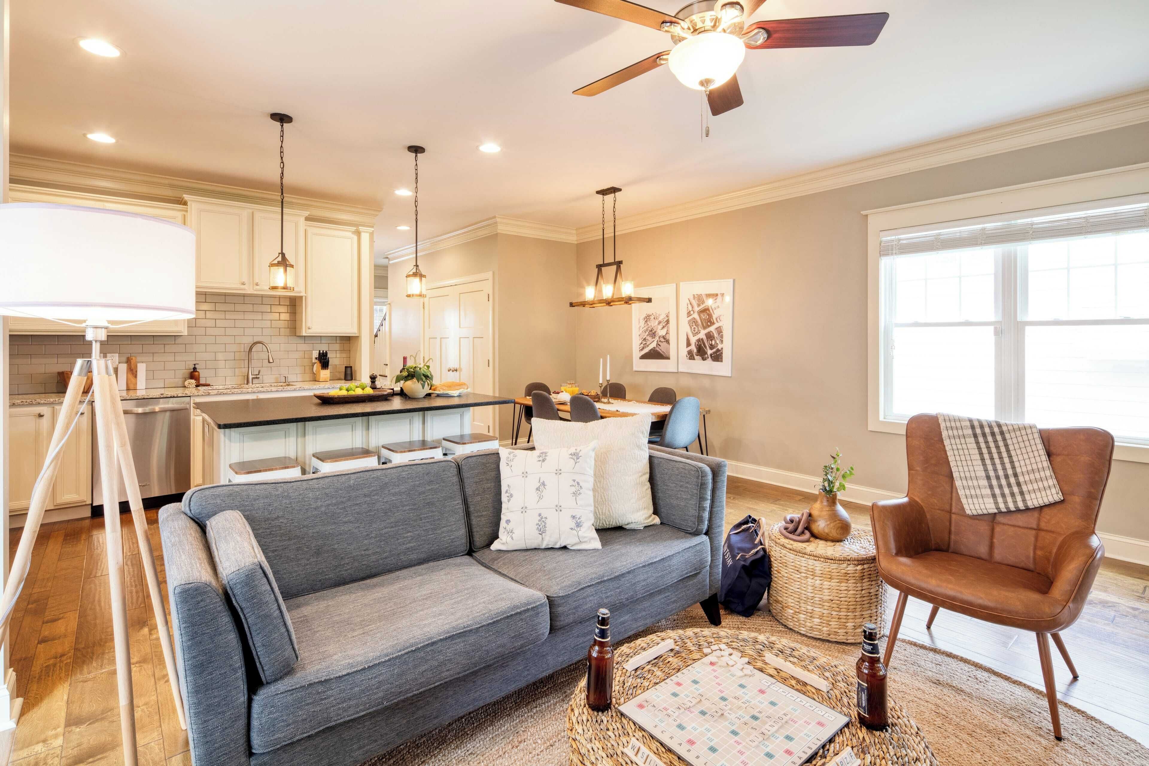 A modern kitchen with white cabinets, a dining area with a table and chairs, a grey sofa, and a brown armchair.