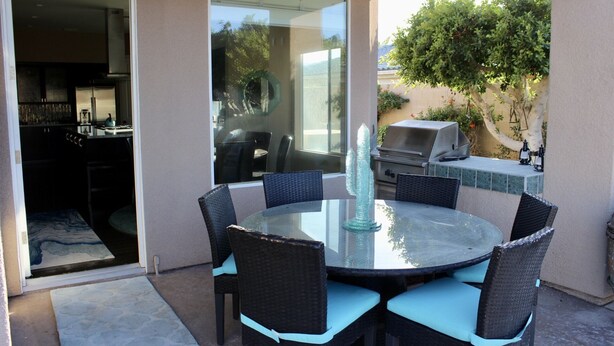 A patio with a round glass table and wicker chairs, a grill, and a view of the backyard. - RANCHO MIRAGE - VICTORIA FALLS ENTERTAINING 4 BD - HUGE POOL & SPA & GRASS YARD! (Rancho Mirage)