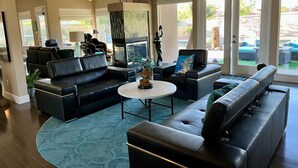 A spacious living room with black leather sofas, a round coffee table, and a fireplace. There are ceiling fans, large windows, and a view of the outdoors. - RANCHO MIRAGE - VICTORIA FALLS ENTERTAINING 4 BD - HUGE POOL & SPA & GRASS YARD! (Rancho Mirage)