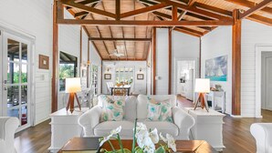 A spacious living area with a high ceiling and wooden beams, featuring a white sofa set, a dining table, and a kitchen in the background.