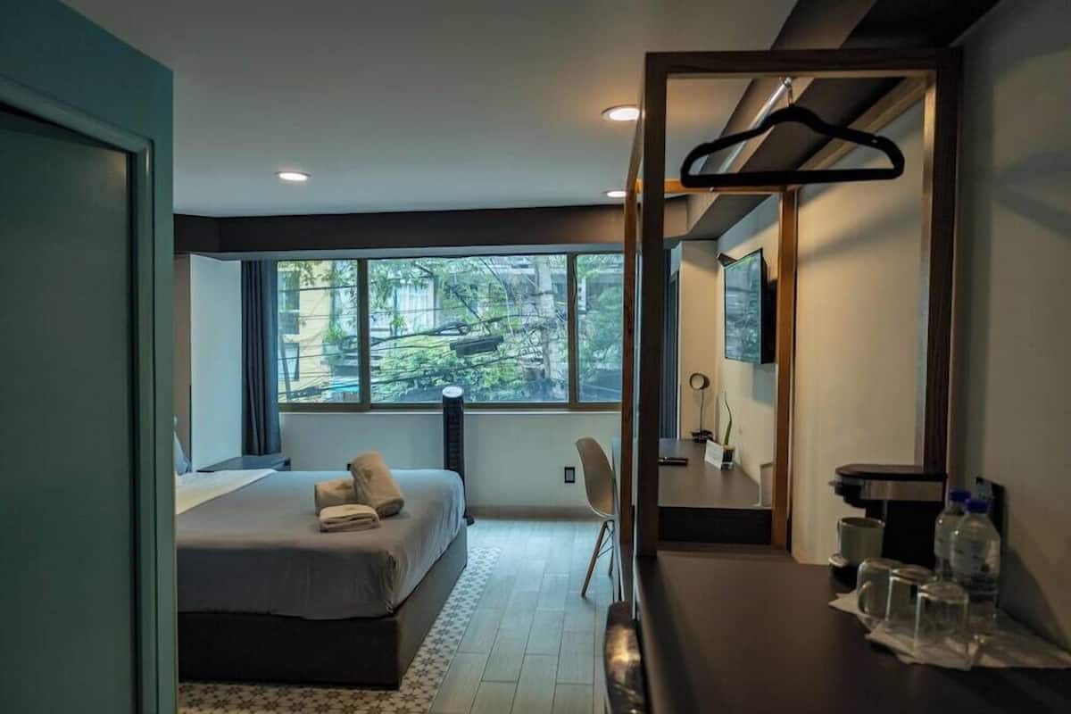 Une chambre d’hôtel moderne, équipée d’un lit, d’un bureau, d’une chaise, d’un miroir et offrant une vue sur la verdure environnante.