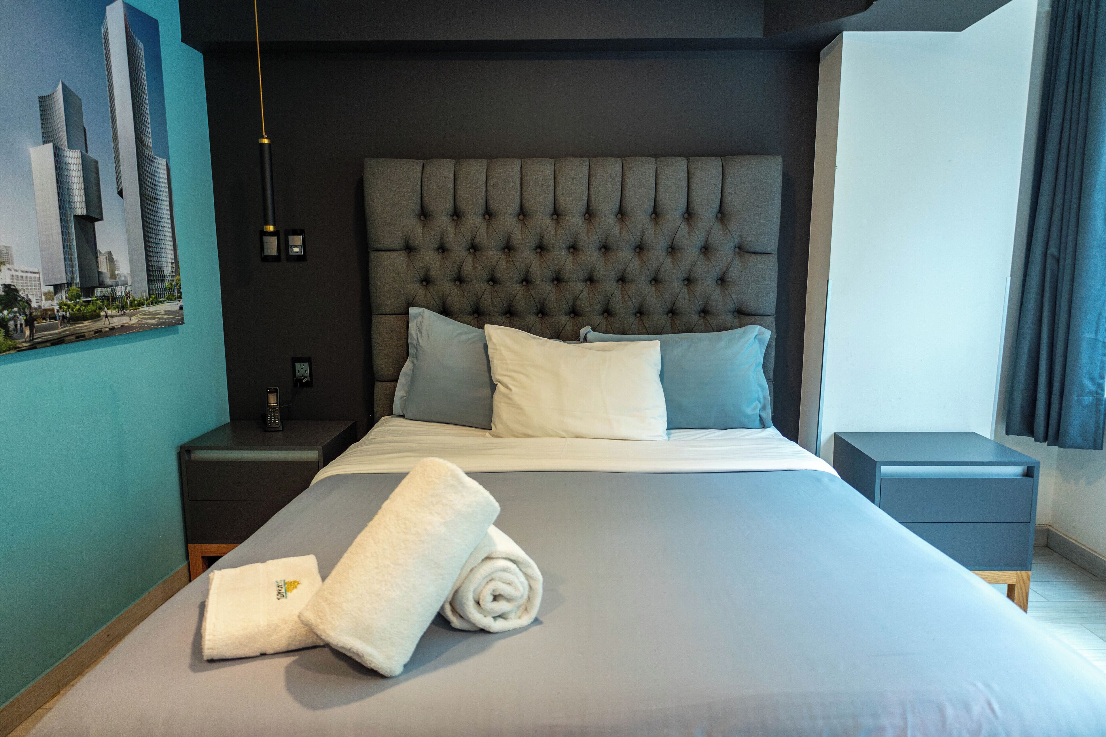A modern hotel room with a large bed, a tufted headboard, and a cityscape artwork on the wall.