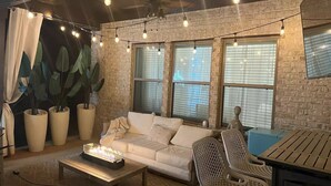 A cozy living area with a white sofa, a coffee table with a lit fireplace, and string lights. There are large potted plants and a ceiling fan. - LUX House! HOT TUB! 4 min to campus. Golf lot (Auburn)