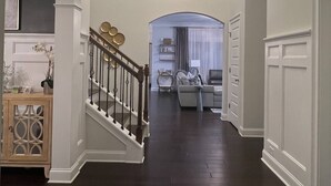 A spacious hallway with a dark wooden floor, white walls, and a chandelier. A staircase with a wooden railing and decorative wall sconces is visible. - LUX House! HOT TUB! 4 min to campus. Golf lot (Auburn)