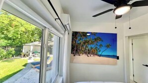 Ein Schlafzimmer mit Bett, Deckenventilator, Fenster mit Gartenblick und Wandgemälde mit Strandmotiv.