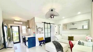 Ein modernes Wohnzimmer mit einem weißen Sofa, einem roten Sessel, einem blauen Tisch und einer Küchenzeile mit Balkonblick.