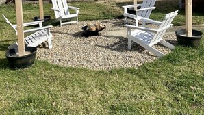 A backyard with four Adirondack chairs arranged around a fire pit, surrounded by a wooden fence and a gravel area. - Perfect for Fall Getaway - Firepit : Walk to Water (Buckeye Lake)