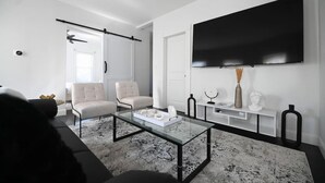 A modern living room with a large flat-screen TV mounted on the wall, a glass coffee table, two armchairs, and a sofa. - 191 Seymour, Ave. Derby · Spacious Renovated 3 bedroom near GriffinHospital (Derby)