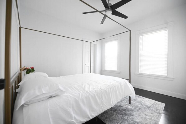Une chambre à coucher avec un lit, un ventilateur de plafond et deux fenêtres équipées de stores.