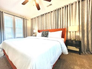 Habitación de hotel con una cama grande, ventilador de techo, mesita de noche con lámpara y ventana con cortinas.