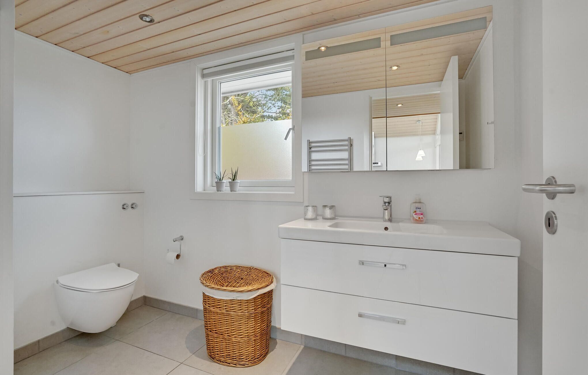 A modern bathroom with a toilet, a wicker basket, a sink with a mirror, and a window with plants.