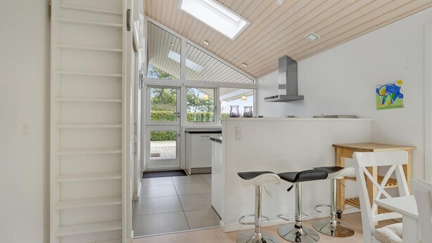 Une cuisine moderne avec des meubles blancs, une cuisinière encastrée et un puits de lumière au-dessus du plan de travail. On y trouve deux tabourets de bar et une table en bois.