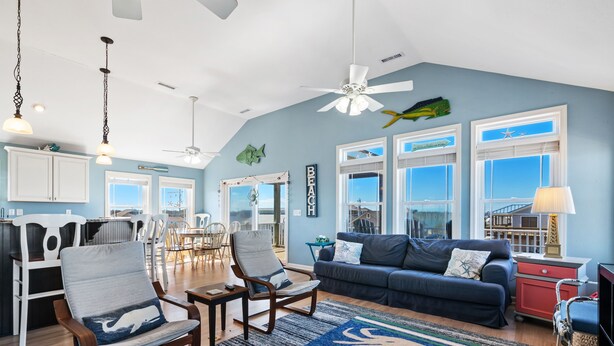 A spacious living area with a blue accent wall, a ceiling fan, and a large window with a view. - Ocean Views with a 1 minute walk to beach access! Linens & Bath Towels included! (Kill Devil Hills)