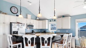 A kitchen with white cabinets, a central island with bar stools, a clock on the wall, and a window with blinds. - Ocean Views with a 1 minute walk to beach access! Linens & Bath Towels included! (Kill Devil Hills)