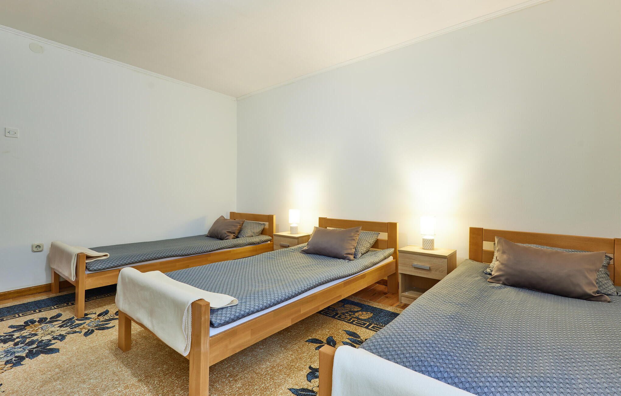 Une chambre avec trois lits simples, chacun doté d’une tête de lit en bois et d’une table de chevet avec une lampe. La chambre est dotée d’un sol recouvert de moquette à motifs floraux.