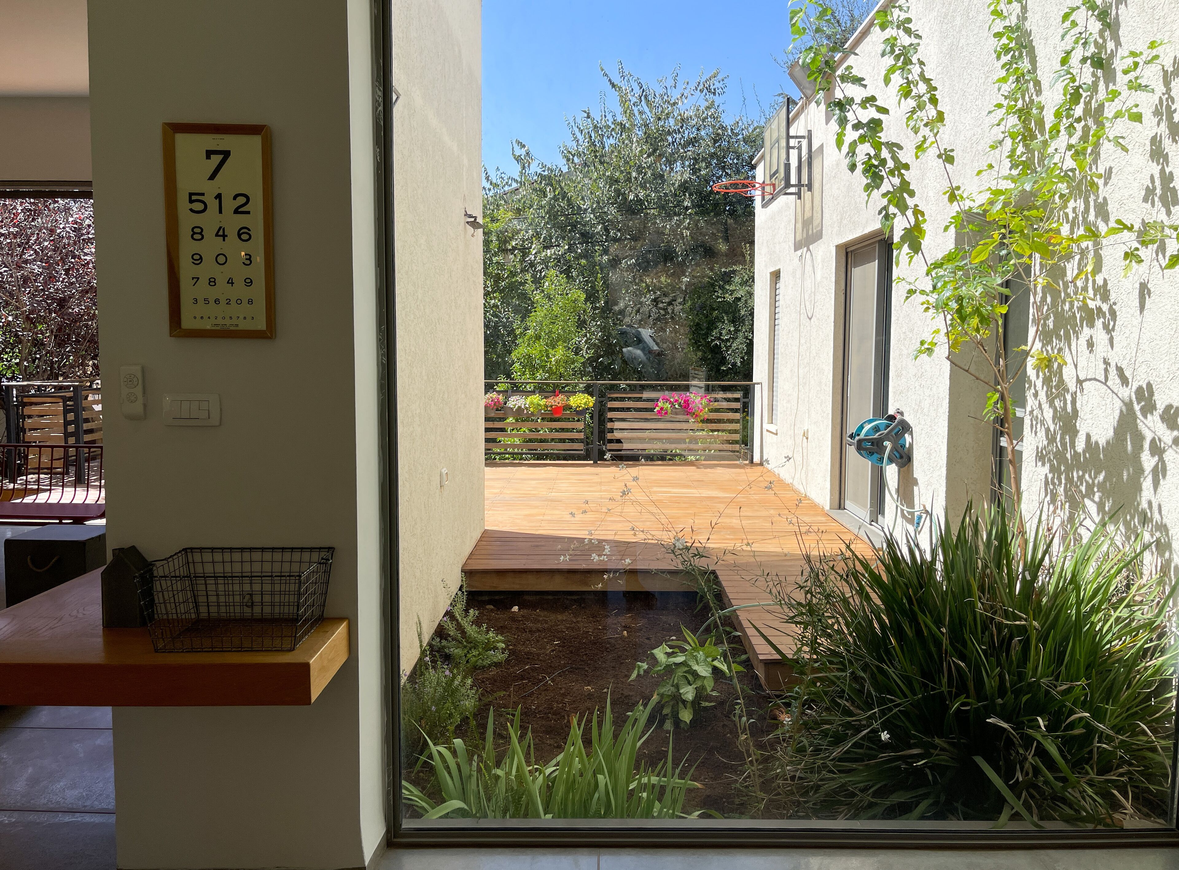 A view through a window to a wooden deck with a metal fence, a bench, and a planter with flowers.