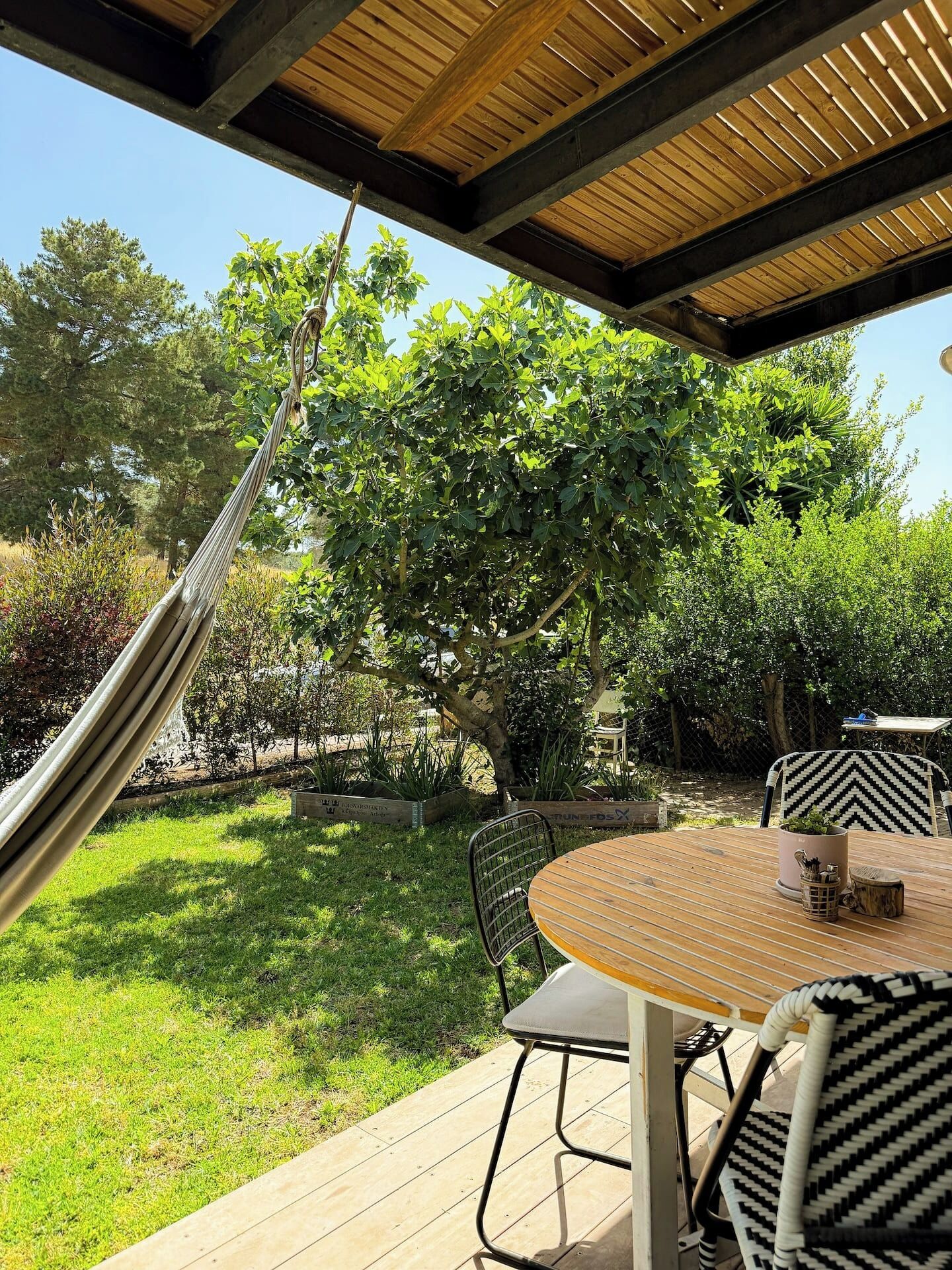 Une terrasse extérieure avec une table ronde en bois, des chaises en métal, un hamac et une vue sur un jardin luxuriant et des arbres.