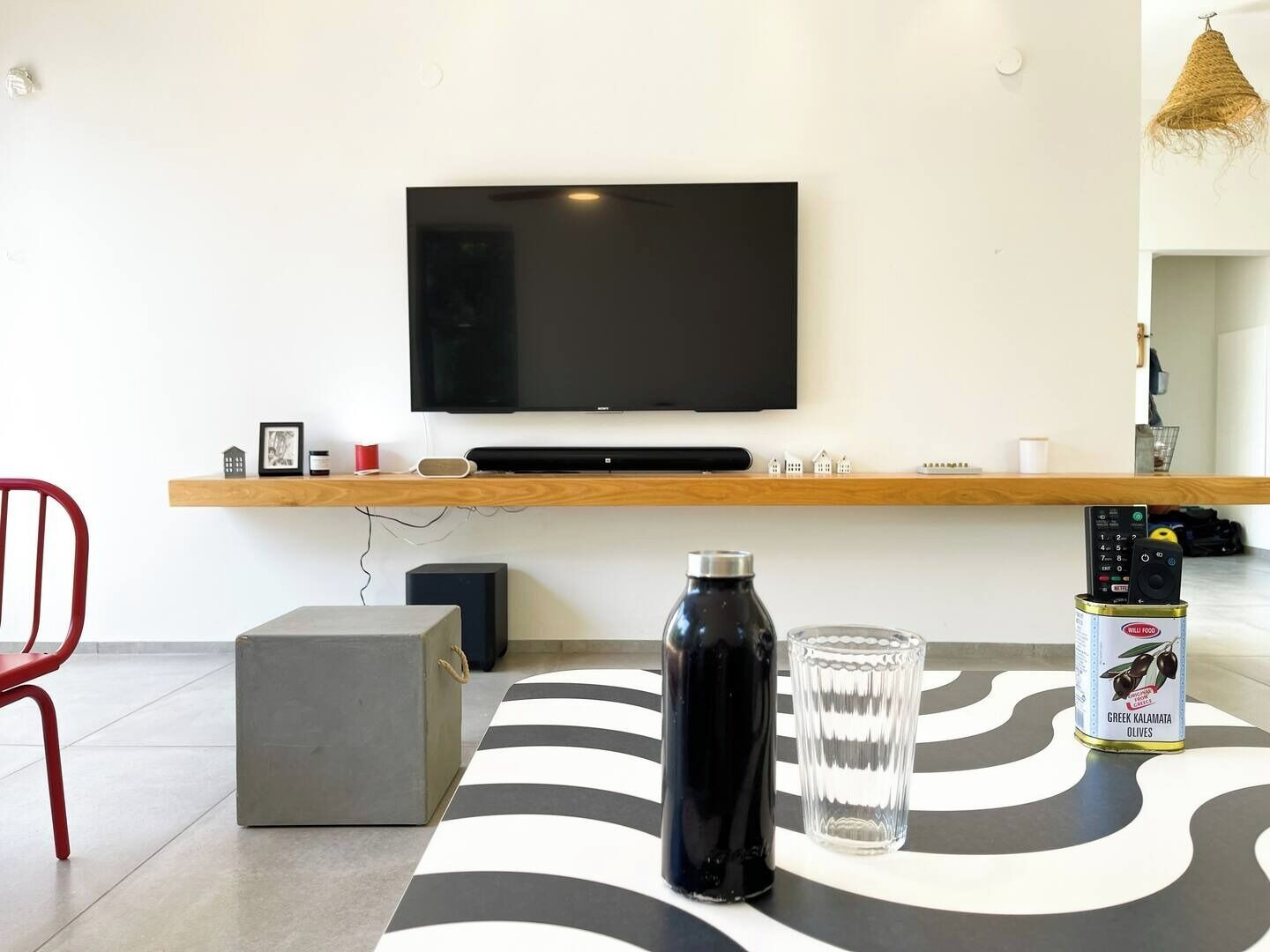A modern living room with a flat-screen TV mounted on a wooden shelf, a black water bottle, a clear glass, and a can of Greek Kalamata olives on a patterned rug.