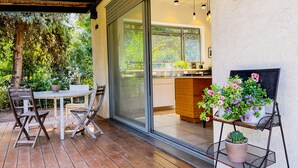 Un patio con una mesa y sillas, una puerta corrediza de vidrio que da a una cocina y plantas en macetas sobre una estantería.